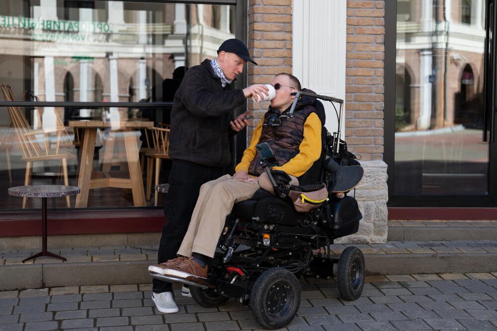 Male caregiver helps man in wheelchair drink from cup.